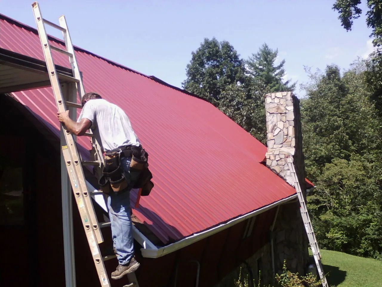 Roofer on a ladder beside a red metal roof and stone chimney — Soffit & Fascia Repair by Roofing Brothers in Hebo