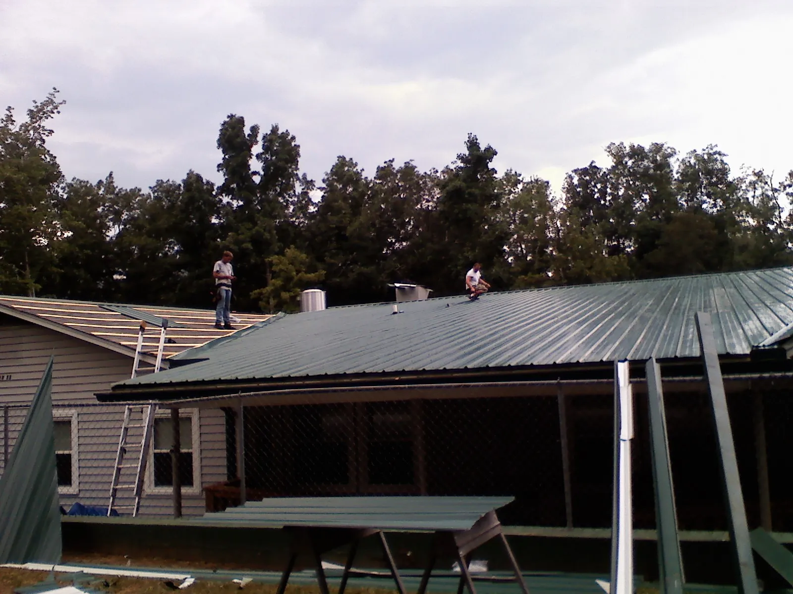 Two roofers installing green metal panels above a covered porch in Hebo